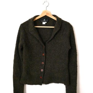 Peut inclure: Cardigan vert olive foncé avec col châle et fermeture boutonnée. Le pull à manches longues est fait d'une matière texturée et est suspendu à un cintre en bois. Les poignets sont repliés. L'étiquette indique "Peruvian Connection".