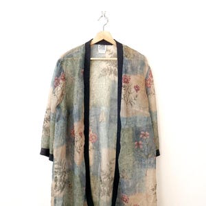 May include: A floral print kimono jacket with a patchwork design. The jacket features a mix of blue, green, and beige squares with pink flower accents. It has black trim along the edges and is hanging on a wooden hanger.