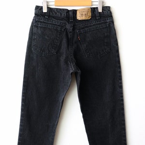 May include: Black denim jeans featuring a high-waisted cut, two back pockets, and a tan leather patch on the waistband with the text Levi Strauss & Co. The jeans include a small orange tab on the right back pocket and are suspended from a hanger.