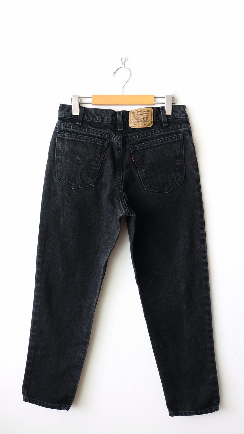 May include: Black denim jeans featuring a high-waisted cut, two back pockets, and a tan leather patch on the waistband with the text Levi Strauss & Co. The jeans include a small orange tab on the right back pocket and are suspended from a hanger.
