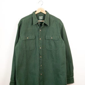 May include: A long-sleeved forest green button-down shirt featuring a pointed collar, a front button placket, and two chest pockets with buttoned flaps. The interior neck label displays the text GS and the size L on a white background.