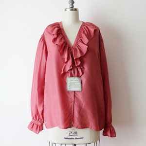 May include: A salmon pink silk blouse with a large ruffled collar and long sleeves. The blouse has a button-up front and a tag that reads "100% Silk".