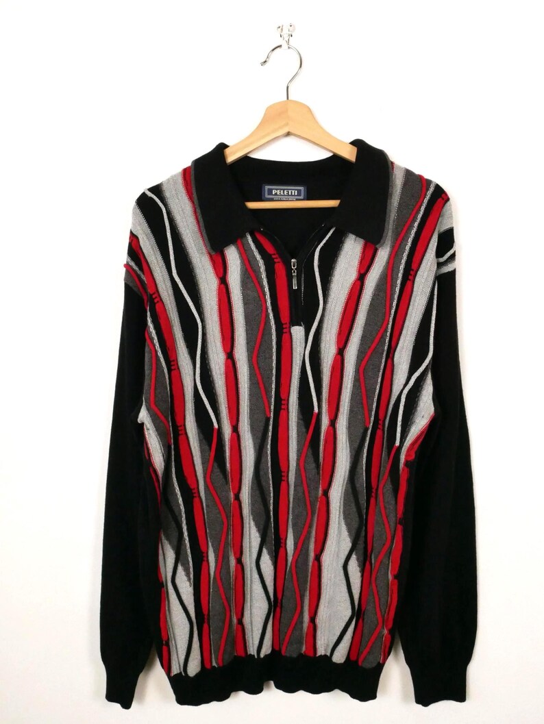 Op de afbeelding: Een gebreid shirt met lange mouwen, een zwarte kraag en een korte ritssluiting aan de voorzijde. Het voorpand heeft een verticaal patroon van golvende rode, grijze en zwarte strepen. Het merklabel Peletti is zichtbaar in de nek.