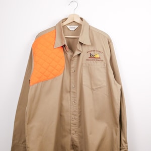Może przedstawiać: Beżowa koszula z długim rękawem z pomarańczową, pikowaną naszywką na ramieniu. Koszula ma zapięcie na guziki, kieszeń na piersi i haftowany napis "Webster County Pheasants Forever". Wisząca na drewnianym wieszaku.