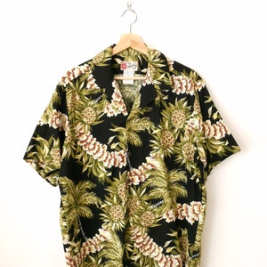 Peut inclure: Une chemise boutonnée à manches courtes noire avec un imprimé ananas et palmier. La chemise a un col et un devant boutonné.