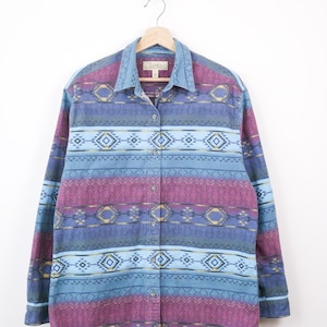 May include: A long-sleeved button-down shirt with a colorful geometric pattern. The shirt features a blue denim collar and cuffs, with a design of blue, purple, and gold geometric shapes. The shirt is hanging on a wooden hanger.