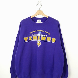 May include: A purple long-sleeve sweatshirt featuring the text National Football League, Minnesota Vikings, and a small Viking helmet graphic in yellow and white. The garment has a crew neckline and a Lee Sport brand label at the inner collar.