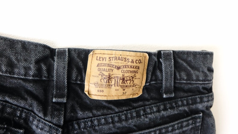 May include: Black denim jeans featuring a tan leather-like patch on the back waistband. The patch displays the text Levi Strauss & Co. Original Riveted Quality Clothing, two horses pulling a pair of jeans, the number 550, and the size W 32.