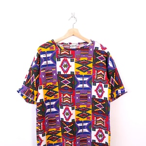 以下が含まれることがあります： 赤、黄、紫、黒の鮮やかな幾何学模様の半袖シャツ。 デザインは、大胆でカラフルなテキスタイルを作成する、繰り返しの形状と線が特徴です。 シャツは白い背景に木製のハンガーに吊るされています。