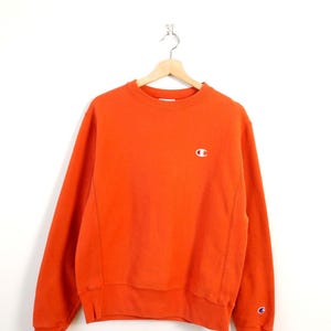 May include: An orange Champion sweatshirt with a small white logo on the chest and a Champion logo on the left sleeve cuff. The sweatshirt is hanging on a wooden hanger against a white background.