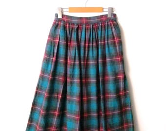 May include: A long, plaid skirt in shades of teal, red, and navy blue hangs from a wooden hanger. The skirt features a classic tartan pattern, with vertical and horizontal lines creating a grid-like design. The fabric appears to be a medium-weight material.
