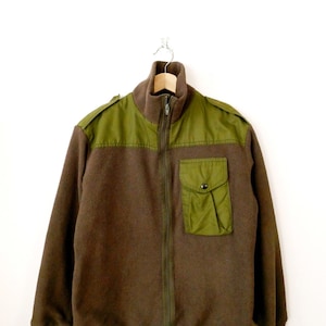 May include: A brown fleece jacket with olive green shoulder patches and a chest pocket. The jacket features a full-length zip and a high collar. The garment is suspended on a wooden hanger against a white backdrop.
