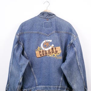 May include: A blue denim jacket with a dark collar and a logo on the back. The logo features a horseshoe, the word "CORRAL", and the words "Restaurant & Saloon" in a western-style design. The jacket is hanging on a wooden hanger.