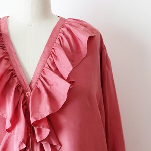 May include: A pink silk blouse with a large ruffled collar and a button closure. The blouse has long sleeves and is made of 100% silk.