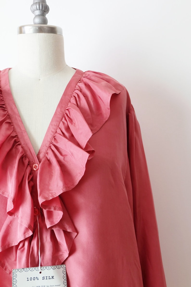 May include: A pink silk blouse with a large ruffled collar and a button closure. The blouse has long sleeves and is made of 100% silk.