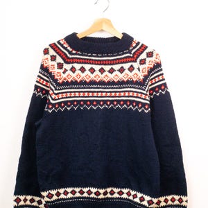 May include: A navy blue long-sleeve jumper featuring a thick ribbed crew neckline and cuffs. The garment displays a horizontal fair isle pattern across the chest and shoulders, incorporating white and red geometric diamond and dot motifs.