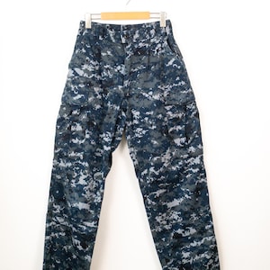 May include: Cargo trousers featuring a blue, grey, and black digital camouflage pattern. These trousers include a button and zip fly closure, belt loops, and two large side cargo pockets with flap closures, all hanging from a wooden hanger.