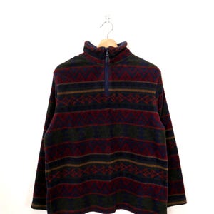 May include: A fleece pullover with a geometric pattern in shades of navy, burgundy, olive green, and brown. The long-sleeved top features a mock neck and a quarter-zip closure. The garment is displayed on a wooden hanger against a white background.