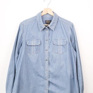 May include: A light blue long-sleeve denim shirt featuring a classic collar, a front button-down closure, and two chest pockets with flaps. The pockets are accented with vertical white stitching details. The shirt is displayed on a wooden hanger.