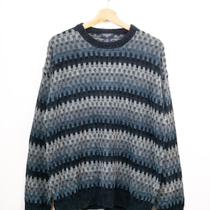 May include: A long-sleeve crew neck sweater featuring a repeating geometric pattern in shades of dark blue, light blue, and grey. The garment has a ribbed knit collar, cuffs, and hem, with a Dockers brand label visible at the inner neckline.