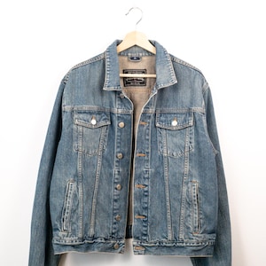 Peut inclure: Une veste en jean bleu délavé moyen avec un col classique, une fermeture boutonnée avec des boutons métalliques, deux poches poitrine à rabat boutonné et deux poches latérales passepoilées. L'étiquette intérieure indique Structure, established 1989.