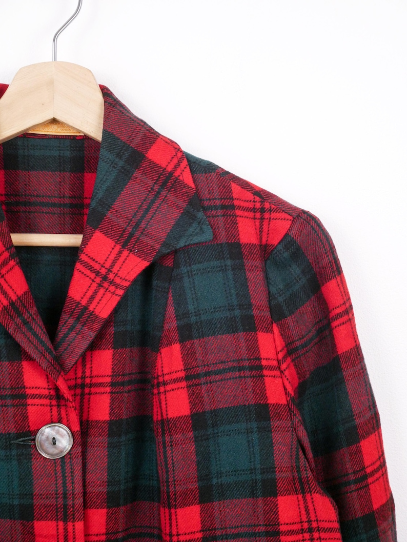 May include: A red and green plaid jacket with a large silver button. The jacket is hanging on a wooden hanger. The plaid pattern is a classic design, with a mix of red, green, and black lines.