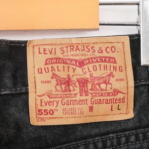 Op de afbeelding: Zwarte spijkerbroek van denim met een beige, leerachtige patch met rode tekst en afbeeldingen. De patch bevat de tekst Levi Strauss & Co. San Francisco, Cal. Original Riveted Quality Clothing, Trade Mark, Patented in U.S. May 20 1873, 550 Relaxed Fit Tapered Leg.