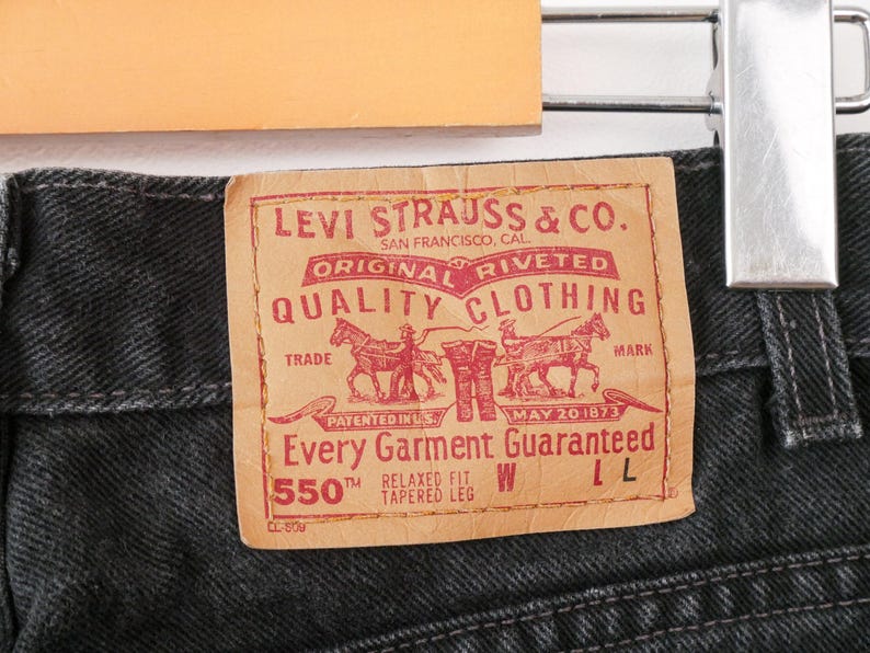 Op de afbeelding: Zwarte spijkerbroek van denim met een beige, leerachtige patch met rode tekst en afbeeldingen. De patch bevat de tekst Levi Strauss & Co. San Francisco, Cal. Original Riveted Quality Clothing, Trade Mark, Patented in U.S. May 20 1873, 550 Relaxed Fit Tapered Leg.