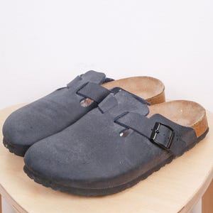 May include: A pair of dark blue clogs with a buckle closure. The clogs have a textured appearance and a cork footbed. The shoes are sitting on a light-colored wooden surface.