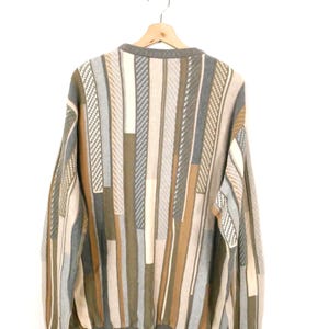 Puede incluir: Un su&eacute;ter multicolor con un patr&oacute;n de rayas verticales en tonos grises, beige y marr&oacute;n. El su&eacute;ter est&aacute; colgado en una percha de madera sobre un fondo blanco. Los pu&ntilde;os y el cuello son de color gris liso.