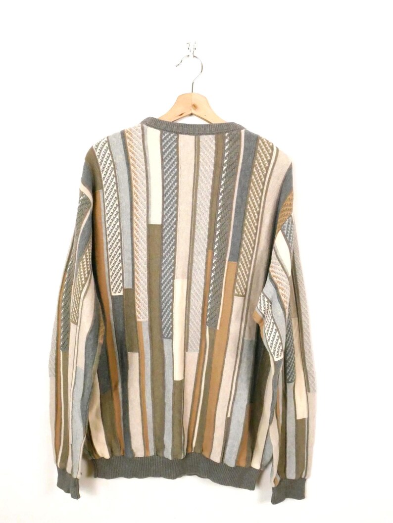 Puede incluir: Un su&eacute;ter multicolor con un patr&oacute;n de rayas verticales en tonos grises, beige y marr&oacute;n. El su&eacute;ter est&aacute; colgado en una percha de madera sobre un fondo blanco. Los pu&ntilde;os y el cuello son de color gris liso.
