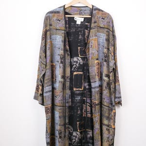 May include: A long, open-front kimono-style jacket with a unique pattern. The jacket features a mix of colors including gray, black, and gold. The design includes geometric shapes and abstract figures. The jacket is hanging on a wooden hanger.