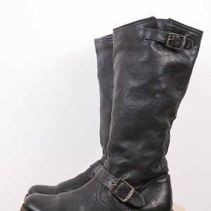 May include: A pair of black leather motorcycle boots with buckle fastenings. The boots have a worn, textured appearance and a mid-calf height. The soles are dark and show signs of wear.