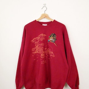 May include: A deep red long-sleeve crewneck sweatshirt featuring an embroidered graphic of a hunting dog and a pheasant. The design includes a printed landscape scene with a tree, a barn, and the word IOWA in a golden-brown color palette.