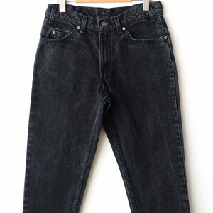 May include: A pair of black denim jeans featuring a high-waisted design, a button and zipper fly closure, five pockets, and belt loops. The jeans are displayed on a wooden hanger with silver metal clips, showcasing a faded wash finish.