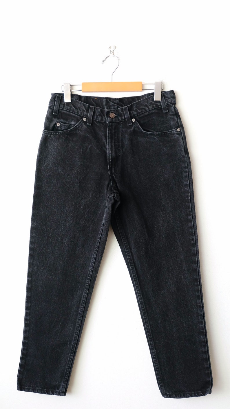 May include: A pair of black denim jeans featuring a high-waisted design, a button and zipper fly closure, five pockets, and belt loops. The jeans are displayed on a wooden hanger with silver metal clips, showcasing a faded wash finish.