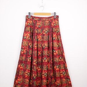 May include: A long, pleated skirt with a vibrant, repeating pattern of fleur-de-lis and diamond shapes in red, orange, yellow, and navy blue. The skirt is hanging on a wooden hanger against a white background.