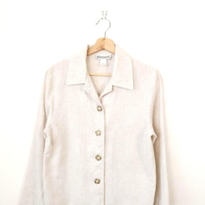 May include: A light beige button-down shirt with long sleeves, hanging on a wooden hanger. The shirt has a collared neckline and several brown buttons down the front. The brand name "BRIGGS" is visible on the label.