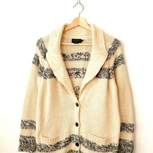 May include: Cream-coloured cardigan sweater with a shawl collar and black and grey striped accents. The sweater has a button-up front, two front pockets, and is hanging on a wooden hanger.