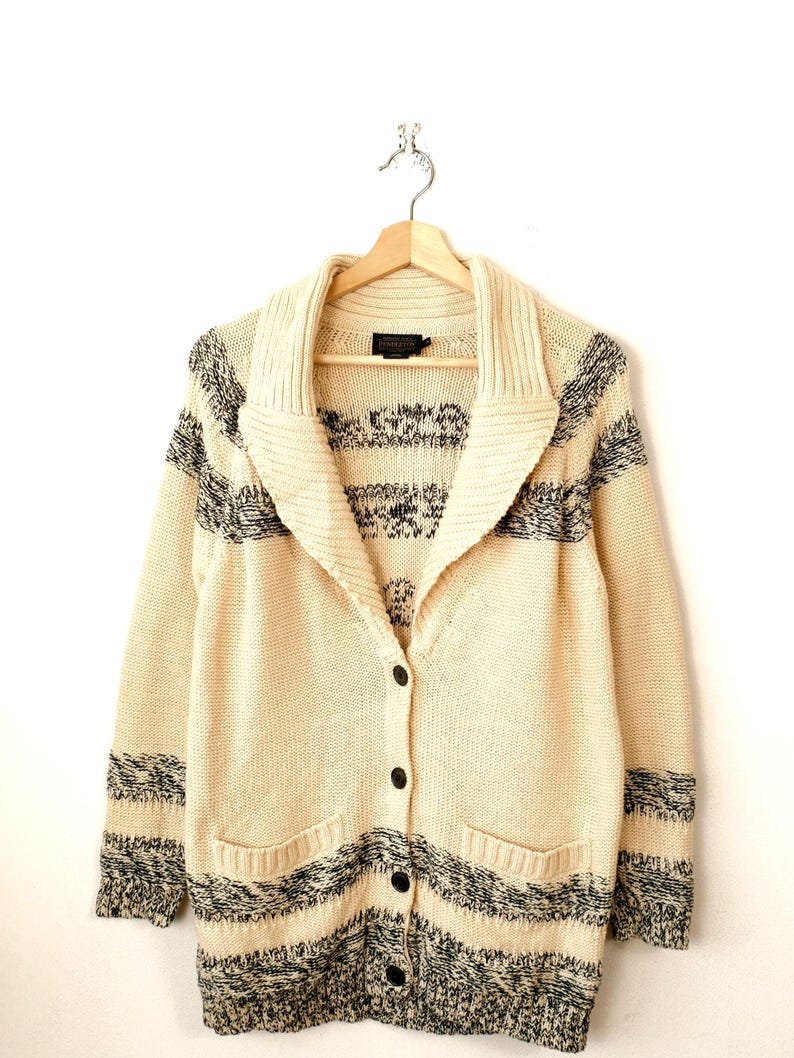 May include: Cream-coloured cardigan sweater with a shawl collar and black and grey striped accents. The sweater has a button-up front, two front pockets, and is hanging on a wooden hanger.