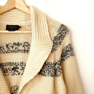 May include: Cream-coloured cardigan sweater with a shawl collar and black geometric patterns. The sweater is hanging on a wooden hanger. The Pendleton label is visible.
