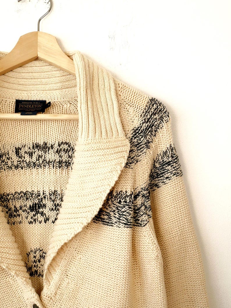 May include: Cream-coloured cardigan sweater with a shawl collar and black geometric patterns. The sweater is hanging on a wooden hanger. The Pendleton label is visible.