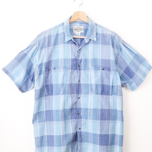 Può includere: Una camicia a maniche corte con bottoni e un motivo a quadri blu e azzurro. La camicia presenta un colletto standard, un'abbottonatura frontale e due tasche sul petto con chiusura a bottone. Un'etichetta del marchio Levi's è visibile sul colletto.