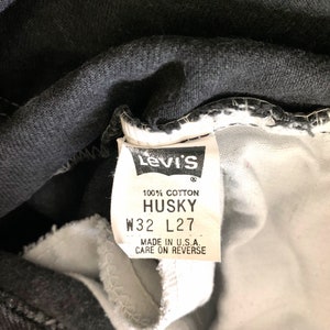 May include: A white fabric clothing tag attached to black denim featuring the Levi's logo, 100% cotton, HUSKY, W32 L27, and the text MADE IN U.S.A. CARE ON REVERSE. The tag is stitched into the interior waistband of the garment.