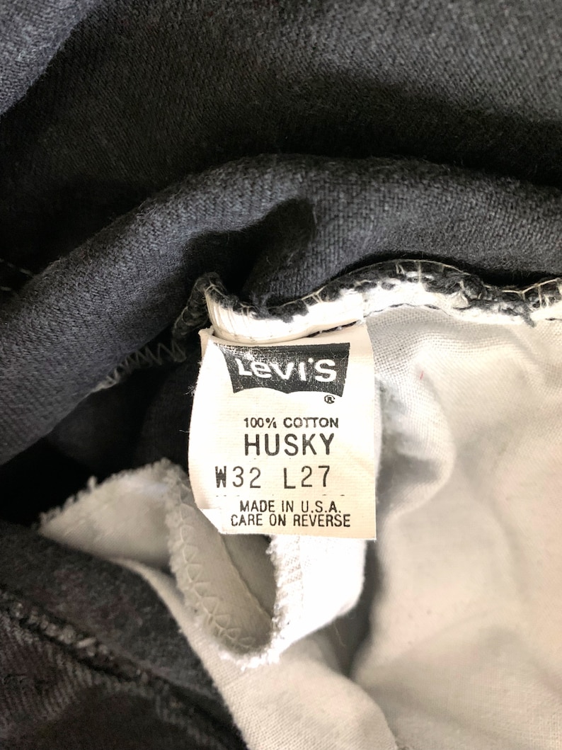 May include: A white fabric clothing tag attached to black denim featuring the Levi's logo, 100% cotton, HUSKY, W32 L27, and the text MADE IN U.S.A. CARE ON REVERSE. The tag is stitched into the interior waistband of the garment.