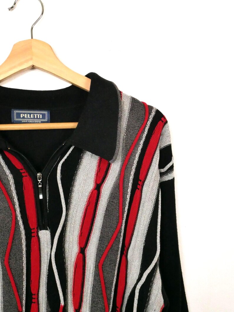 Op de afbeelding: Een zwart, rood en grijs gebreide trui met een kwartritssluiting en een omgeslagen kraag. Het kledingstuk heeft een gestructureerd verticaal streeppatroon in afwisselende kleuren. Een blauw label met de tekst PELETTI is in de nek genaaid.