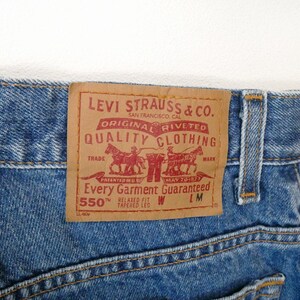 Może przedstawiać: Dżinsy z niebieskiego denimu z brązową sk&oacute;rzaną naszywką Levi Strauss & Co., na kt&oacute;rej widnieje napis "Original riveted quality clothing" i "Every garment guaranteed 550&trade; Relaxed fit tapered leg W LM".