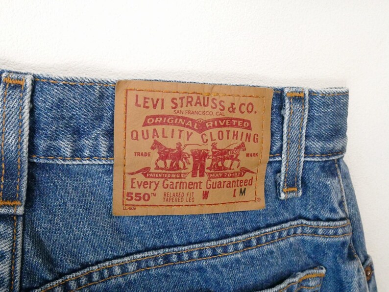 Może przedstawiać: Dżinsy z niebieskiego denimu z brązową sk&oacute;rzaną naszywką Levi Strauss & Co., na kt&oacute;rej widnieje napis "Original riveted quality clothing" i "Every garment guaranteed 550&trade; Relaxed fit tapered leg W LM".