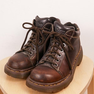 May include: A pair of dark brown leather ankle boots featuring a rounded toe, thick brown laces, and silver-toned metal eyelets. The boots have a thick, dark brown rubber sole with visible yellow stitching along the welt and the Dr. Martens logo.