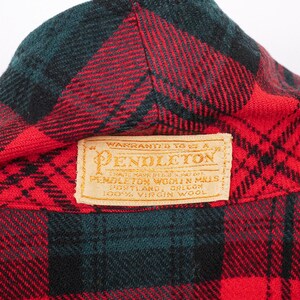 May include: Close-up of a red, black, and green plaid wool fabric. The fabric has a Pendleton label with text that reads "Warranted to be a Pendleton" and "100% Virgin Wool".